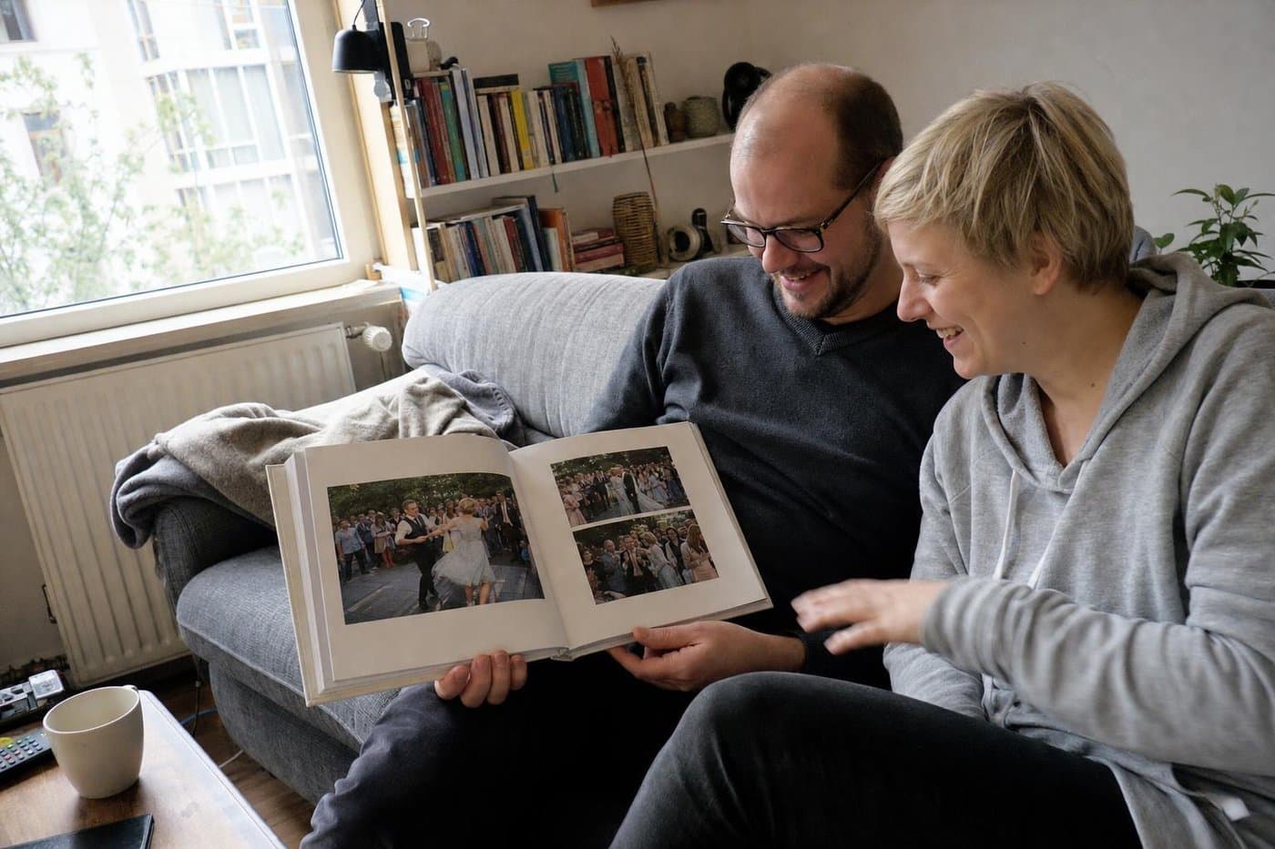 Paar sitzt im Wohnzimmer auf dem Sofa und blättert lachend durch sein Hochzeitsfotobuch