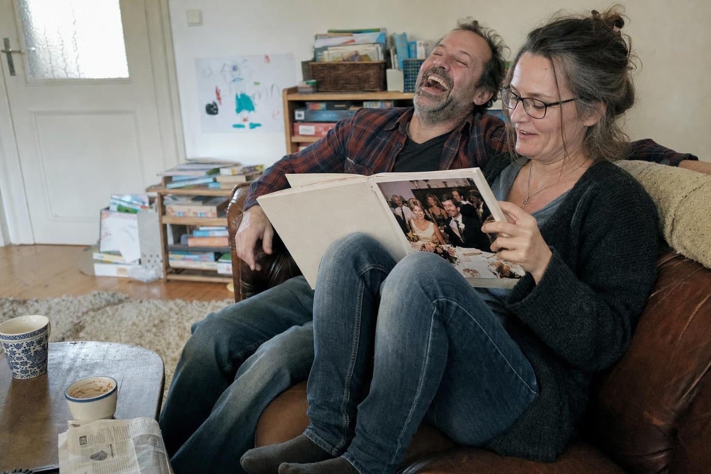 Paar lacht am Sonntagmorgen auf dem Ledersofa über ein Bild in seinem Hochzeitsfotobuch