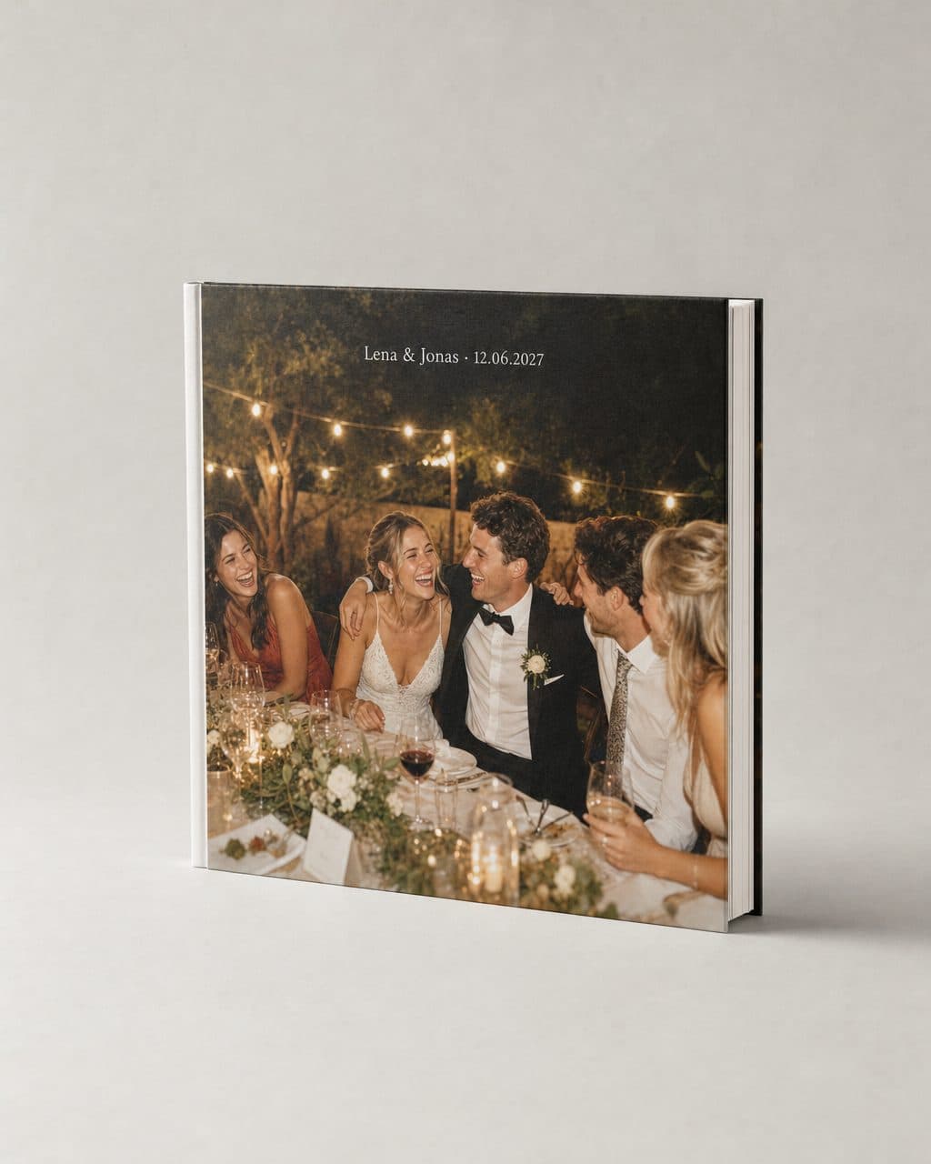 Quadratisches Hardcover Fotobuch, auf dem Cover lacht das Brautpaar mit Freunden an der Gartentafel unter Lichterketten